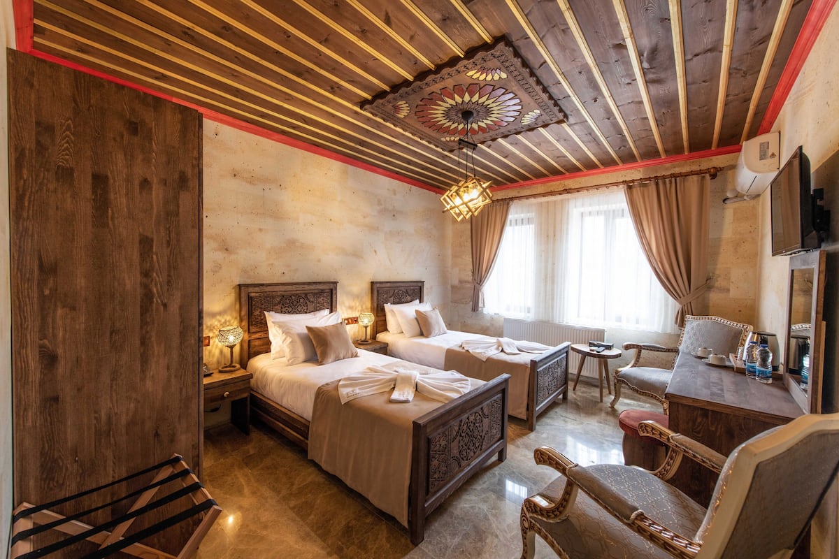 Quarto de hotel com duas camas, cabeceiras de madeira, um candelabro, uma escrivaninha com cadeira e uma televisão.