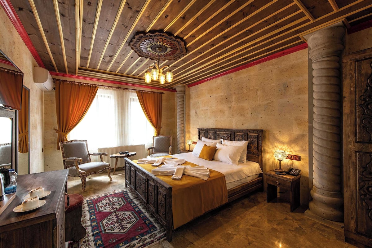 Quarto de hotel com teto de madeira, uma cama grande com cabeceira, uma escrivaninha com cadeira, uma mesinha de apoio com abajur e um tapete estampado.