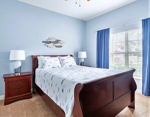 A bedroom with a wooden bed, two bedside tables with lamps, a wall-mounted fish decoration, and a window with blue curtains. - Disney Getaway Oasis: 4BR Townhome, Heated Pool & BBQ, Perfect for Families! (Kissimmee)