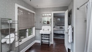 Un bagno con un lavabo, una finestra con persiane, un angolo doccia e un letto visibile sullo sfondo.