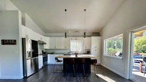 A modern kitchen with a central island, white cabinetry, stainless steel appliances, and a large window providing a view of the outdoors. - Spectacle Lake Waterfront Retreat Chalet (Tonasket)