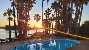 A wooden deck with a swimming pool, surrounded by palm trees and a view of the sunset over a body of water.