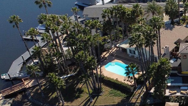 An aerial view of a resort with a swimming pool surrounded by palm trees, a boat dock, and a large building with a sloped roof.
