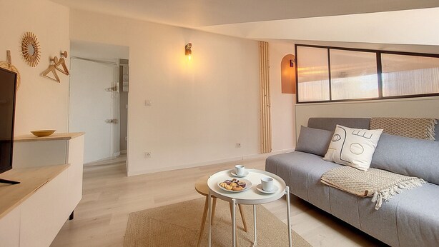 A modern living room with a grey sofa, a small round table with a tray of snacks, a television on a stand, and a decorative wall piece. - N&J -  VIEUX NICE SUITE - Old Town - Close sea (Niza)