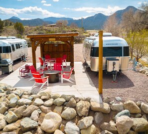 Un camping con vehículos recreativos, un área de picnic techada con sillas rojas y una parrilla.
