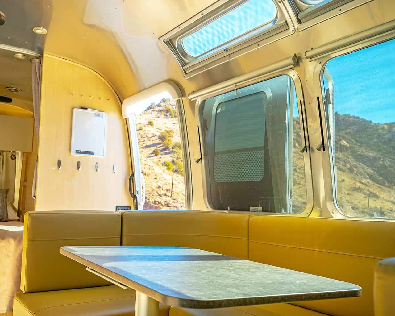 A vehicle interior with a table, cushioned seating, a window with a view of a desert landscape, and a control panel on the wall.