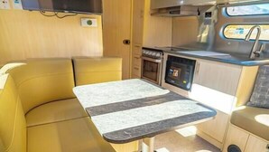 Ein kompakter Küchenbereich mit Tisch und Sitzgelegenheit, einem an der Wand befestigten Fernseher und einem Fenster, das für natürliches Licht sorgt.