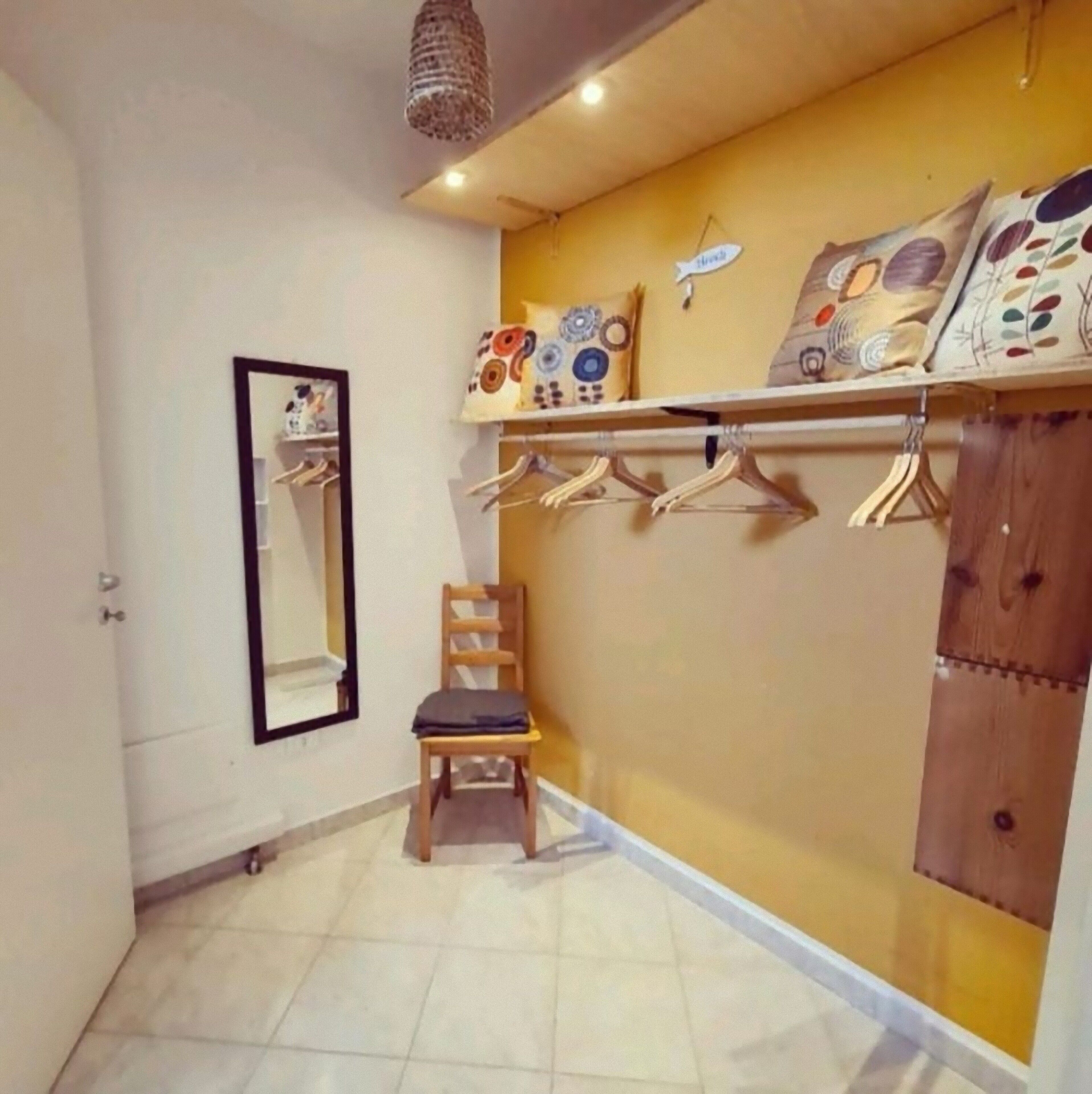 Een kleine, goed verlichte kamer met een betegelde vloer, een houten stoel, een spiegel, een plank met hangende kleding en een houten garderobe.