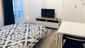 A bedroom with a bed, a TV on a wooden stand, a white desk, and a window with curtains.