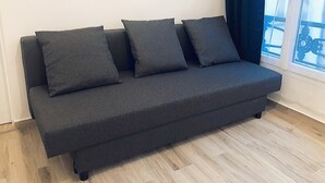 A dark grey sofa with four cushions against a white wall, next to a window with a blue curtain.