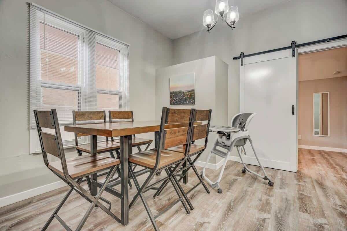A dining area with a wooden table and metal chairs, a white desk with a chair, and a cityscape painting on the wall.