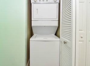 A stacked washer and dryer unit in a corner with a door and a window with blinds.