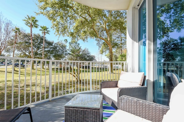 A balcony with wicker furniture, a glass table, and a view of a grassy area with trees.
