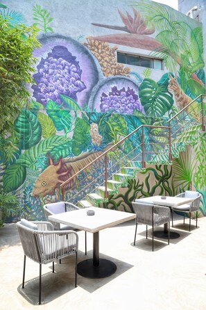 Une terrasse extérieure ornée d’une fresque représentant une scène de la préhistoire, avec un dinosaure et une végétation luxuriante. On y trouve trois tables et quatre chaises.