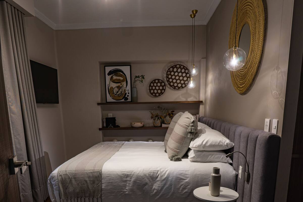 Une chambre à coucher moderne avec un grand lit, des tables de chevet, un miroir rond et des œuvres d’art décoratives au mur.