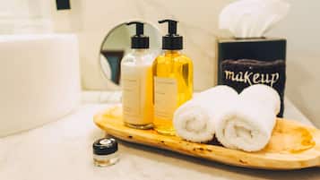 Ein Badezimmer-Waschtisch mit einer Holzschale, die zwei Lotionflaschen, eine kleine Handdesinfektionsmittel-Flasche und zwei gerollte Handtücher enthält.