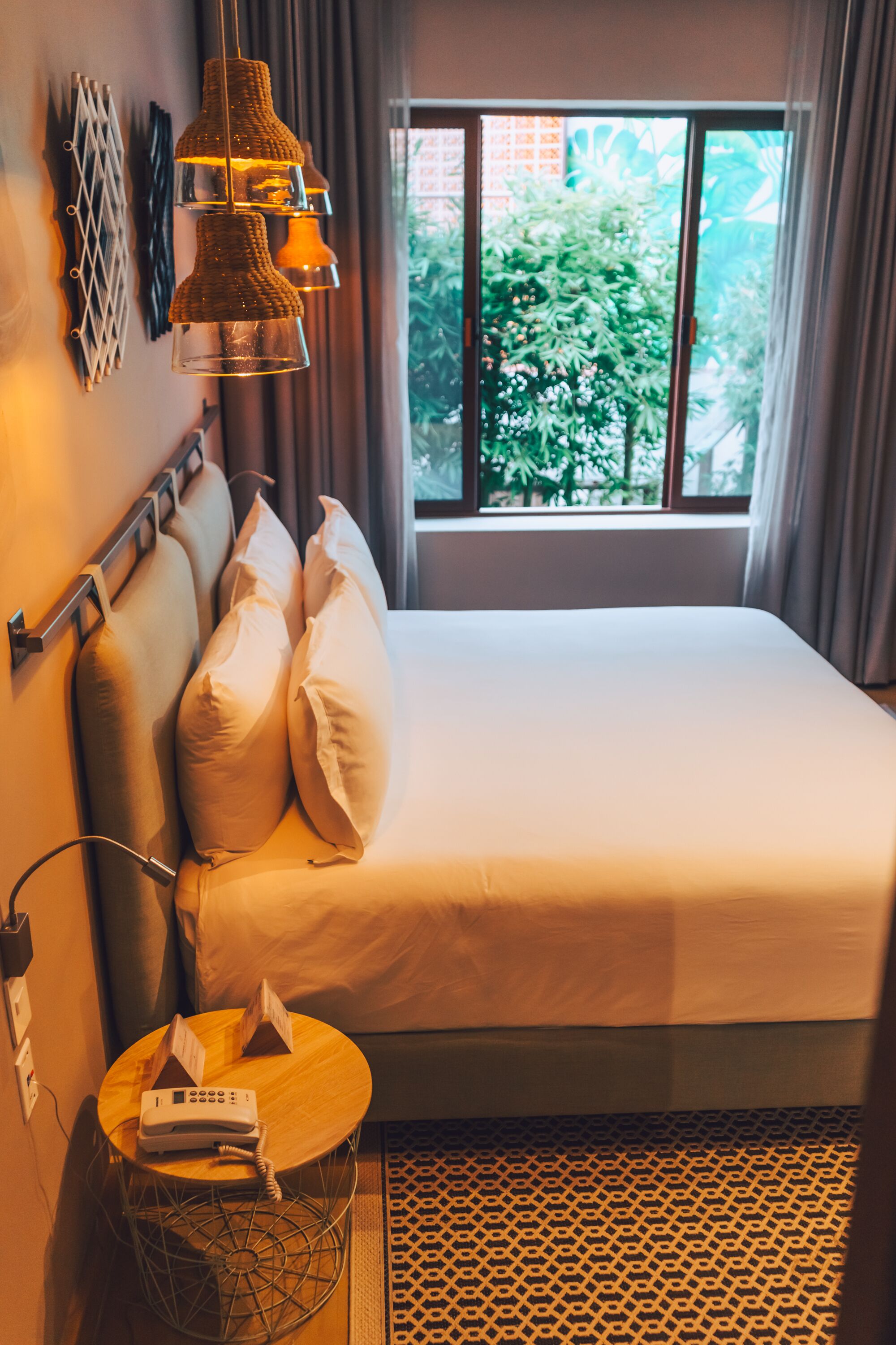 Ein Hotelzimmer mit Bett, Kissen, einem Nachttisch mit Telefon, einer Wanddekoration und einem Fenster mit Blick auf Grünflächen.