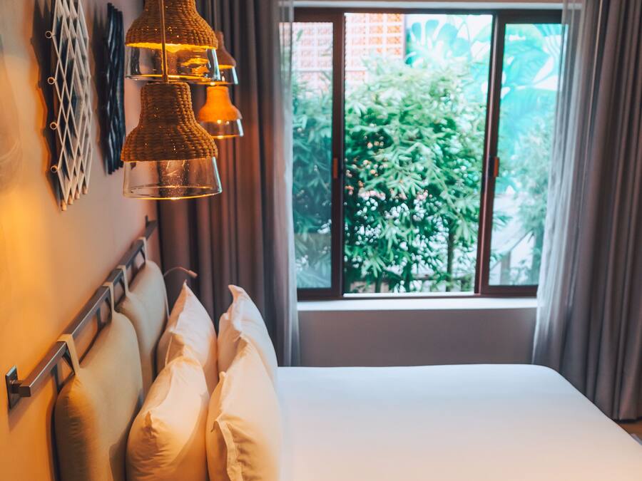 Ein Hotelzimmer mit Bett, Kissen, einem Nachttisch mit Telefon, einer Wanddekoration und einem Fenster mit Blick auf Grünflächen.