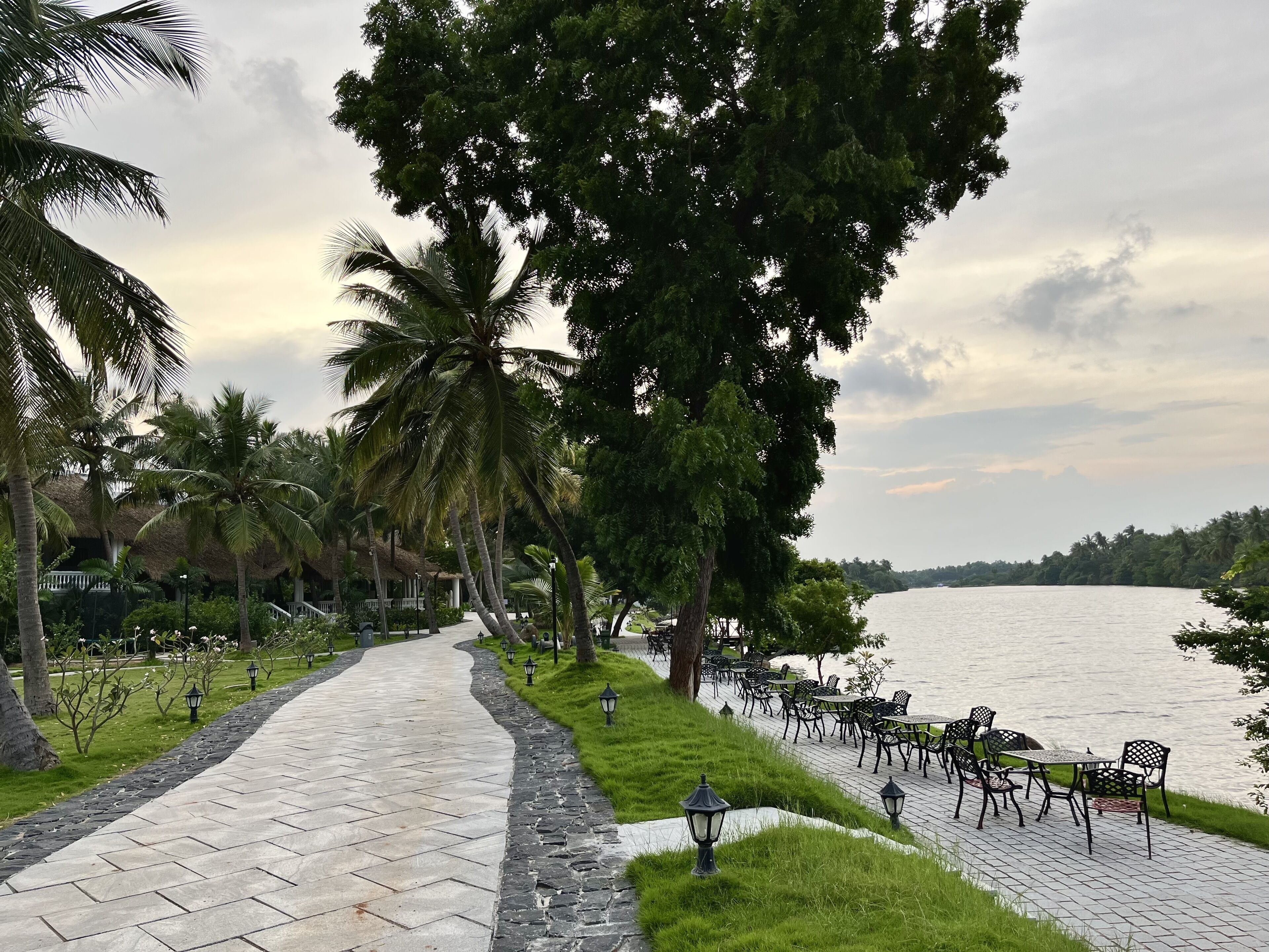 A paved pathway leading to a riverside seating area with tables and chairs, surrounded by palm trees and greenery.