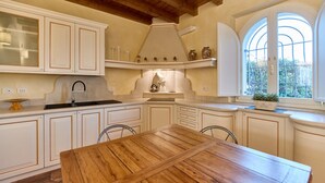 A kitchen with white cabinetry, a wooden dining table, and a window with a view. - Villa Ardea Exclusive Holigarda (Soiano del Lago)
