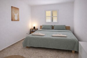 Une chambre à coucher comprenant un lit, des oreillers, une table de chevet avec une lampe, une fenêtre avec des stores et une œuvre d’art accrochée au mur.