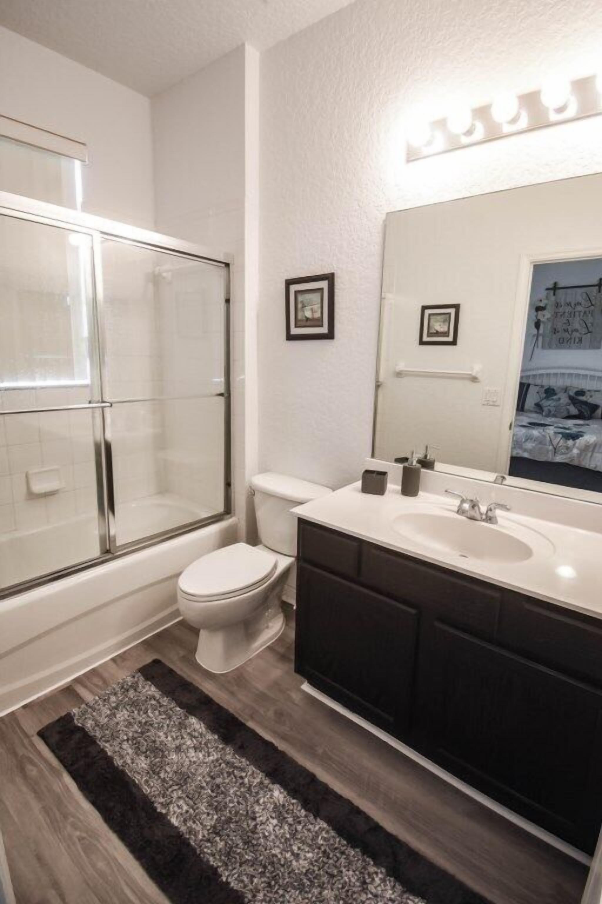 Et badeværelse med glasvæg omkring brusebad, toilet, vaskebord med spejl og en måtte på gulvet.