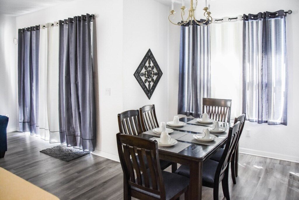 A dining room with a wooden table set for four, flanked by curtains and a chandelier.