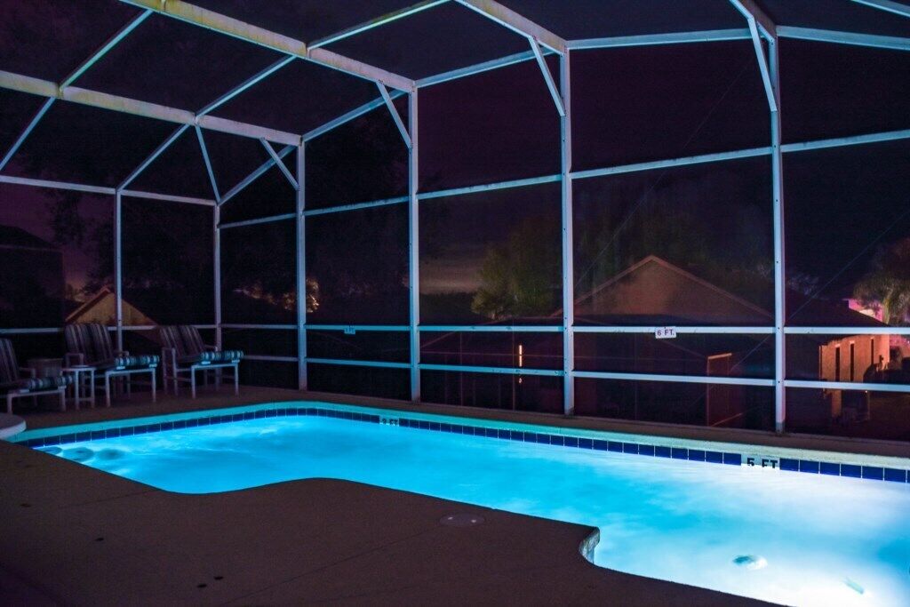 Una piscina con cerramiento de vidrio por la noche, rodeada de sillones de descanso.