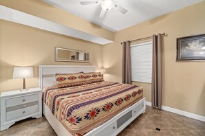 Une chambre à coucher comprenant un lit, une table de chevet, une lampe, un ventilateur de plafond, une fenêtre avec des rideaux et un tableau encadré accroché au mur.