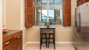 A kitchen with a window, wooden shutters, a stainless steel refrigerator, and a small table with two stools.