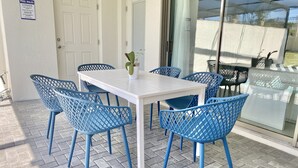 Une terrasse avec une table blanche et des chaises bleues, une porte et une porte vitrée donnant sur un espace abrité.