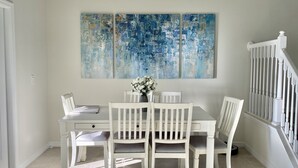Un coin repas avec une table et des chaises blanches, un vase de fleurs et un grand tableau abstrait accroché au mur.
