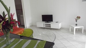 Ett modernt, minimalistiskt vardagsrum med en platt-TV på ett vitt skåp, ett litet vitt bord med en krukväxt och en grön och orange stol.