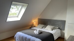 A bedroom with a sloped ceiling, a skylight, a bed with white and grey bedding, a bedside table with a lamp, and a radiator. - Maison Familiale " L'abbé Garo " (Port-en-Bessin-Huppain)