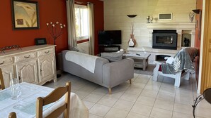 A living room with a sofa, TV, fireplace, and dining area with a table and chairs. - Maison Familiale " L'abbé Garo " (Port-en-Bessin-Huppain)
