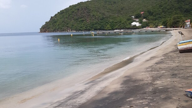A beach with a sandy shore, a pier extending into the water, and a hill with buildings in the background. - Balaou Villa facing the sea (anse d'arlet)