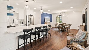 Une cuisine moderne avec des meubles blancs, un îlot central doté de tabourets de bar, un coin repas avec une table et des chaises en bois, et un fauteuil en osier avec un coussin.