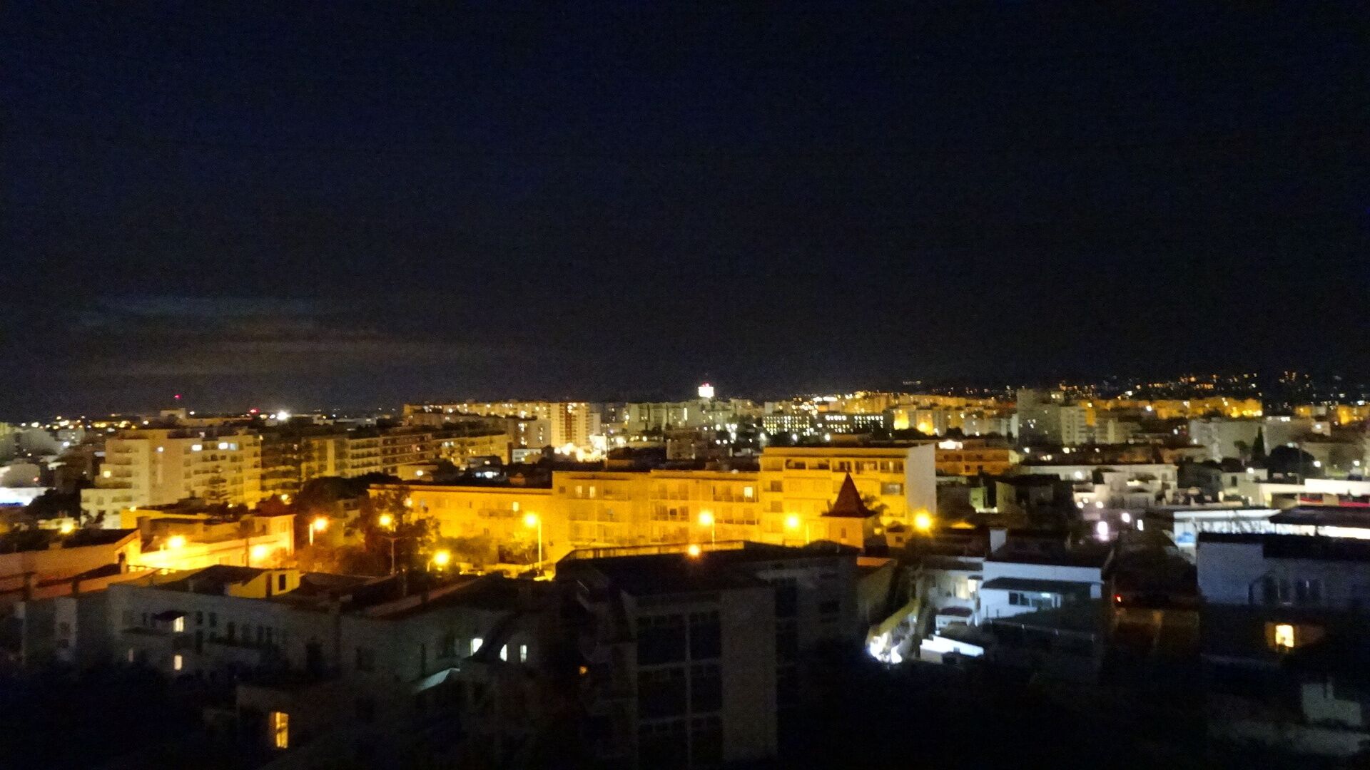 Une vue nocturne d’une ville, avec des bâtiments illuminés et une imposante structure fortifiée au premier plan.
