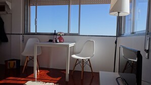 Una habitación pequeña y moderna con un escritorio blanco, dos sillas blancas, un florero rojo con flores y un ventanal con vistas al cielo.