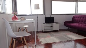 Una sala de estar con un escritorio blanco, un sofá morado, un televisor sobre un mueble bajo y un ventanal grande.
