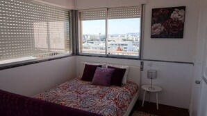 Un dormitorio con cama, ventana con persianas, vista a la ciudad y un cuadro en la pared.