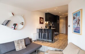 Een moderne woonkamer met een grijze bank, een klein rond wit schap met boeken en een teddybeer, en een transparante glazen tafel met stoelen.
