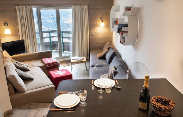 A modern living room with a sofa, a dining table set for two, and a view of a mountainous landscape through large windows.