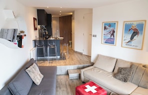 A modern living room with a grey sofa, beige sofa, and a red Swiss flag cushion. There is a kitchen area with a bar and two clear chairs. The room has wooden flooring and features two posters on the wall.