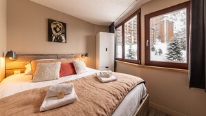 Habitación de hotel con una cama grande, una mesita de noche con lámpara, un armario y una ventana que ofrece vistas a árboles nevados y edificios.