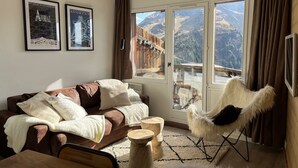 A cozy living room with a brown sofa, white cushions, a white furry chair, and a wooden coffee table. There are two framed posters on the wall and a large window with a mountain view. - Immodreams - Le Dahu - Avoriaz (Avoriaz)