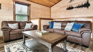 Un interior de cabaña acogedor con dos sofás de cuero marrón, una mesa de centro de madera y un ventanal con persianas.