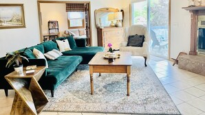 Een woonkamer met een groene bank, een houten salontafel en een witte fauteuil. Er is een open haard, een raam en een deur naar buiten.