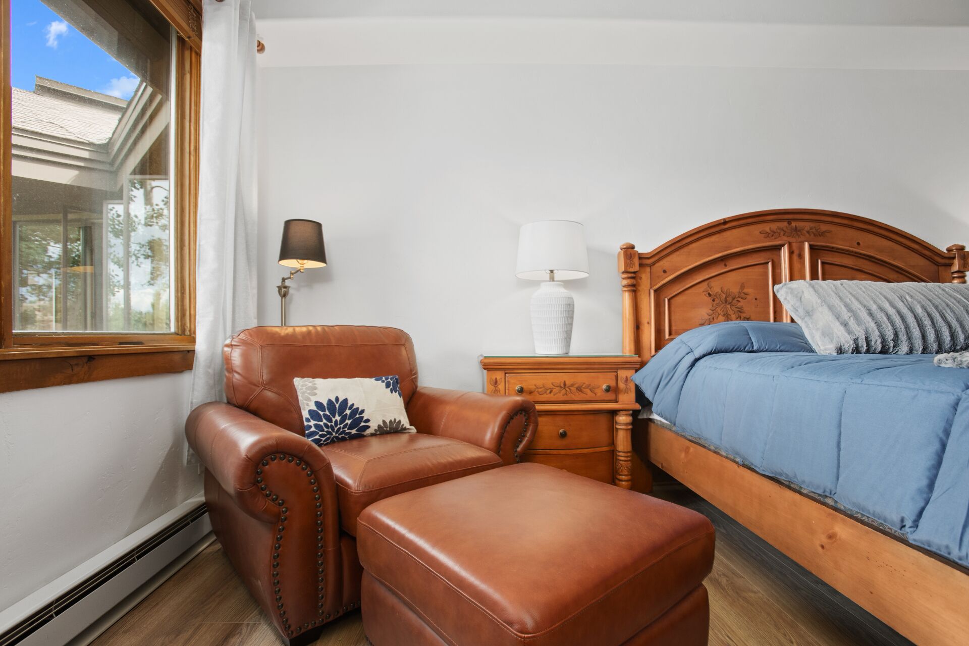 Una camera da letto con una poltrona in pelle, un letto di legno, un comodino con lampade e una finestra vista alberi.