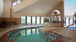 Una piscina interior con vistas al exterior, un área de descanso con sillas y una estructura de piedra con plantas.